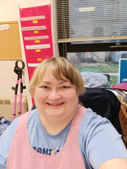 A woman sits at a desk and takes a joyful selfie. Behind her, files are organized on a hanging folder and pink crutches lean against the wall.